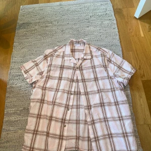 Rutig kortärmad skjorta från ASOS DESIGN - Säljer en snygg kortärmad skjorta från ASOS DESIGN i ljusrosa och brunt rutigt mönster. Skjortan har klassisk krage och knäppning framtill. Perfekt för en avslappnad och trendig stil.