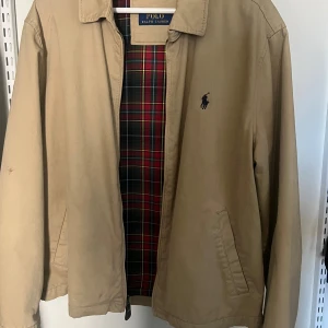 Beige jacka från Polo Ralph Lauren - Säljer en klassisk beige jacka från Polo Ralph Lauren med rutig insida i rött, grönt och gult. Jackan har krage, dragkedja och två snedställda fickor framtill. Ikoniska broderade loggan på bröstet. Perfekt för vår och höst! Liten fläck på ena ärmen, se sista bilden. Utöver det i super fint skick ✨