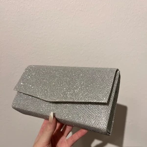 Glittrig silverfärgad kuvertväska - glittrig kuvertväska i silver från Boohoo. Kommer med en kedja så man kan bära den över axeln också.🩷