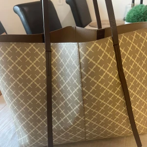 Beige mönstrad Malene Birger shoppingväska med bruna handtag - I fint skick! Säljer en stor beige shoppingväska med vitt rutmönster och långa bruna handtag. Väskan är rymlig och har en matchande innerficka. Perfekt för dig som vill ha plats med allt du behöver under dagen.