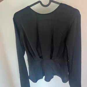 Svart omlottblus med lång ärm - Säljer en svart omlottblus med lång ärm och markerad midja. Blusen har djup v-ringning i ryggen och ett lätt glansigt tyg som ger en elegant känsla. Perfekt att styla med jeans eller kjol. Använt fåtal gånger i storlek S🤍
