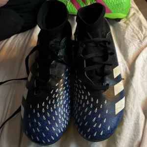 Adidas blå fotbollsskor med vita detaljer - Snygga blå fotbollsskor från Adidas med vita ränder på sidorna och vita droppformade detaljer över tån. Skorna har svart snörning, högre strumpliknande skaft och gula dobbar undertill. Perfekta för dig som vill sticka ut på planen.