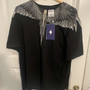 Svart t-shirt Marcelo Burlon med vingar - Svart t-shirt från Marcelo Burlon County of Milan med ikoniskt vingtryck i grått och vitt runt halsen. Klassisk passform och korta ärmar, tillverkad i mjuk bomull. Perfekt statement-plagg för dig som gillar streetwear.