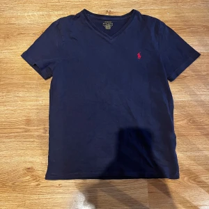 Mörkblå t-shirt från Polo Ralph Lauren - Snygg mörkblå t-shirt från Polo Ralph Lauren med röd broderad logga på bröstet. Modellen har v-ringning och korta ärmar, samt en slim fit passform. Tillverkad i mjuk bomull som känns skön mot huden. Perfekt för en clean och stilren look.