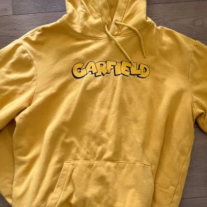 Gul Garfield hoodie från HM oversized - Säljer en gul hoodie med Garfield-tryck på bröstet. Tröjan har huva med dragsko och en stor magficka. Perfekt för dig som gillar bekväma och färgglada plagg med en rolig twist.