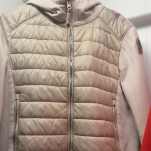 Para jumpers beige Cardigan - Säljer en beige quiltad jacka från Parajumpers med huva och dragkedja framtill. Jackan har lång ärm och är tillverkad i fleece och syntetmaterial. Perfekt för kyliga dagar och har en sportig look.