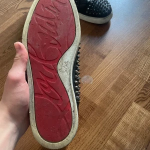 Svarta sneakers med nitar från Christian Louboutin - Säljer ett par svarta sneakers från Christian Louboutin med ikonisk röd sula och silverfärgade nitar över hela skon. Skorna har snörning och är tillverkade i skinn. Perfekta för dig som vill sticka ut med din stil. Skriv om det skulle vara något 🍾