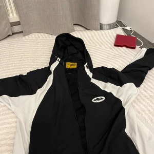 Corteiz Windbreaker - Snygg svart och vit vindjacka från Guerilla med huva och dragkedja. Jackan har sportig look, meshfoder på insidan och coolt logomärke på bröstet. Perfekt för dig som gillar streetwear och vill ha en lätt jacka med stilren design.