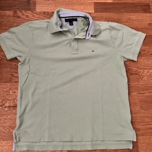 Grön pikétröja från Tommy Hilfiger - Snygg ljusgrön piké från Tommy Hilfiger med klassisk krage och korta ärmar. Tröjan har en liten broderad logga på bröstet och är tillverkad i mjuk bomull. Perfekt för en clean och stilren look.