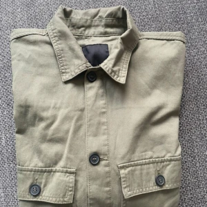 Grön overshirt med fickor och knappar stl L - Stilren grön  overshirt med klassisk krage, två bröstfickor med knappar och raka ärmar. Jackan har en avslappnad passform och är tillverkad i bomull, perfekt att slänga på över en hoodie eller t-shirt. Snygga svarta knappar framtill och på ärmsluten.