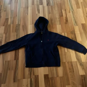 Marinblå hoodie från Polo Ralph Lauren - Snygg marinblå hoodie från Polo Ralph Lauren med dragkedja och huva. Klassisk röd logga broderad på bröstet och två fickor framtill. Perfekt för en chill och stilren look. Passar dig som gillar enkel och tidlös streetstyle.