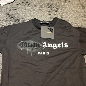 Svart Palm Angels t-shirt Paris print - Svart t-shirt från Palm Angels med stort Paris-print framtill i vitt och svart, där loggan har en cool spray paint-effekt. Klassisk rund hals och korta ärmar. Tillverkad i mjuk bomull, perfekt för en streetwear-look.