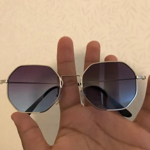 Silvriga åttakantiga solglasögon - Snygga solglasögon med åttakantiga glas och tunn silvrig metallbåge. Glasen har en mörk toning som går från lila till blått, vilket ger en cool och modern vibe. Perfekta för att sticka ut i mängden och ge din stil en extra edge.
