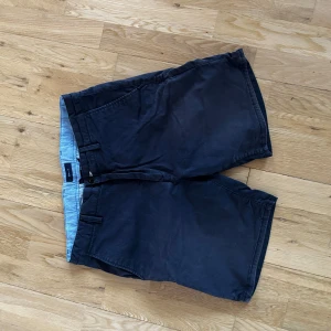 Mörkblå shorts från Gant - Klassiska mörkblå shorts från Gant med snygg passform och diskreta fickor på sidorna. Tillverkade i mjuk bomull med ljusblå insida och randiga detaljer vid linningen. Perfekta för varma dagar och enkel att matcha med t-shirt eller skjorta.