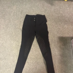 Svarta högmidjade ridbyxor med grip - Snygga svarta ridbyxor med hög midja och silikongrip på insidan av benen för extra grepp. Byxorna har smal passform, bälteshällor och dekorativa bakfickor med små knappar. Perfekta för dig som vill ha stil och funktion i stallet. Köpte för 1099! Är xs men jag som själv har s kan ha på med dessa