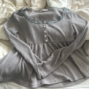 Grå ribbad topp med spets från Romwe - Supersöt grå ribbad topp från Romwe med lång ärm, knappar framtill och spetskant runt halsen. Modellen är lite utsvängd nertill och har en feminin vibe. Perfekt att styla med jeans eller kjol för en chill look. Har ett litet hål se bilden 
