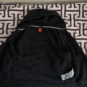 Svart Nike tach med huva M - Svart vindjacka från Nike i storlek M. Jackan har huva, dragkedjor på fickorna och en vit detalj längs insidan av kragen. Tillverkad i ett lätt syntetmaterial som passar perfekt för blåsiga dagar. Enkel och stilren design med klassisk Nike-logga på insidan.år lite skadad under armen men syns inte så mycket 