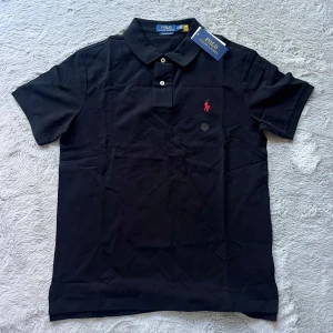 Svart pikétröja från Polo Ralph Lauren - Klassisk svart pikétröja från Polo Ralph Lauren med röd broderad logga på bröstet. Tröjan har korta ärmar, krage och två knappar framtill. Modellen är custom slim fit och passar perfekt till en stilren look.