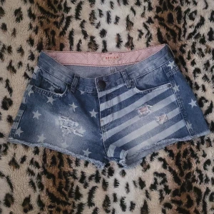 Blå jeansshorts med stjärnor och ränder - Snygga blå jeansshorts i storlek s från Review med coolt mönster av stjärnor och ränder, inspirerat av amerikanska flaggan. Shortsen har slitna detaljer, råa kanter och klassisk femficksmodell. Perfekta för sommaren☀️