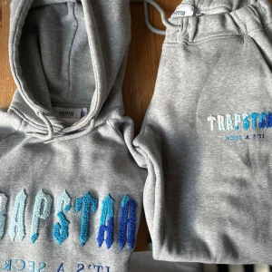Grått Set Trapstar hoodie + byxor med blå brodyr - Säljer ett grått set hoodie+byxor från Trapstar (ÄKTA) med stor broderad logga i olika blå nyanser på bröstet. Fint skick. XS