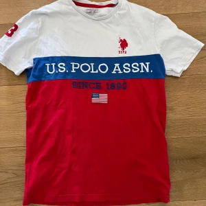 Flerfärgad t-shirt från U.S. Polo Assn. - Säljer en snygg t-shirt från U.S. Polo Assn. i vitt, blått och rött med tryck och broderad logga framtill. T-shirten har korta ärmar och rund halsringning med röd kant. Perfekt för en sportig look!