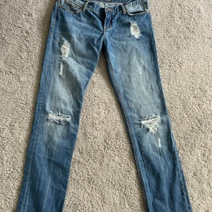 Blå slitna jeans från Hollister - Säljer ett par blå jeans från Hollister med slitna detaljer och låg midja. Jeansen har klassisk femficksdesign, bootcut ben och snygga slitningar framtill för en trendig look.
