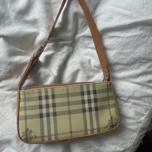 Beige axelväska - Säljer en beige axelväska med klassiskt rutigt mönster och bruna skinnliknande detaljer. Väskan har dragkedja upptill. Perfekt storlek för det viktigaste och stilren design.