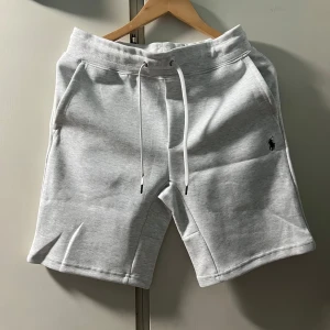 Grå shorts från Polo Ralph Lauren - Snygga ljusgrå shorts från Polo Ralph Lauren med broderad logga på vänster ben. Modellen är slim fit med snörning i midjan och praktiska sidofickor samt en bakficka. Perfekta för en avslappnad stil.