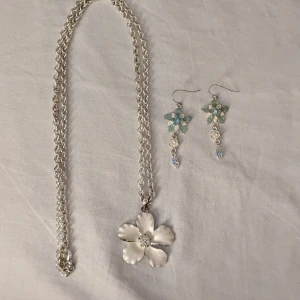 Blommigt halsband och örhängen i silverfärg - Säljer ett set med halsband och örhängen. Halsbandet har en stor vit blomma som hänge och örhängena har blommor i ljusblått och vitt med små pärldetaljer. Perfekt för dig som gillar blommiga smycken och vill ha något unikt.