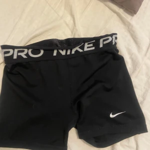 Svarta Nike Pro shorts - Svarta tighta träningsshorts från Nike Pro med bred resår i midjan där det står 'NIKE PRO' i vitt. Perfekta för gymmet eller löpning. Klassisk Nike-logga på ena benet.