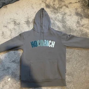 Grå hoodie från Hoodrich - Säljer en stilren grå hoodie från Hoodrich med broderad text i blå och vit på framsidan. Den har en klassisk känguruficka och justerbar huva med snören. Perfekt för en avslappnad stil.