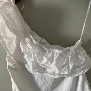 Vit volang topp med spetsdetaljer - Säljer en söt vit volang topp med vackra spetsdetaljer och volang vid axelbanden. Perfekt för en somrig look. Toppen har en ribbad struktur som ger en fin passform.