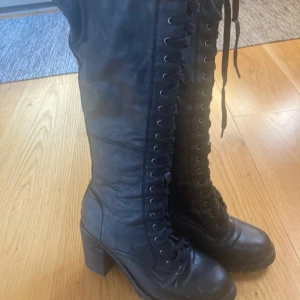Svarta höga boots med snörning - Snygga svarta höga gotiska boots i skinn med snörning och dragkedja. De har en robust klack och en rutig röd och svart insida. Perfekta för en cool look!