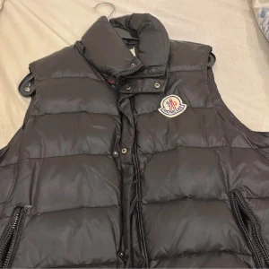 Svart dunväst från Moncler - Säljer en snygg svart dunväst från Moncler med dragkedja och knappar. Perfekt för kyligare dagar och ger en stilren look. Västen har Monclers logga på bröstet och är både praktisk och trendig.