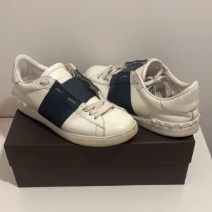 Valentino Open mörkblå - Snygga vita Valentino Garavani sneakers med en bred mörkblå rem över ovansidan. Skorna har snörning och en klassisk design som passar perfekt för en stilren look.