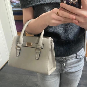 Beige handväska med gulddetaljer - Snygg beige/grå handväska med stilrena gulddetaljer och praktisk dragkedja. Perfekt vardags väska man får plats med det mesta. 