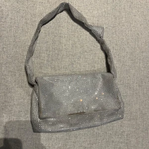 Grå glittrig axelväska  - Snygg grå axelväska med glittriga detaljer från Gina tricot. Väskan har en praktisk magnetknäppning och ett innerfack med dragkedja för extra säkerhet. Perfekt för att ge din outfit lite extra bling!