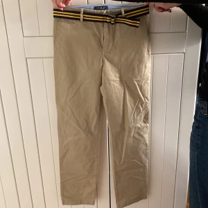 Beige chinos från Polo Ralph Lauren - Snygga beige chinos från Polo Ralph Lauren med ett stilrent bälte i blått och gult. Byxorna har en klassisk design med raka ben och två bakfickor med knappar. Perfekta för en avslappnad men ändå uppklädd stil.