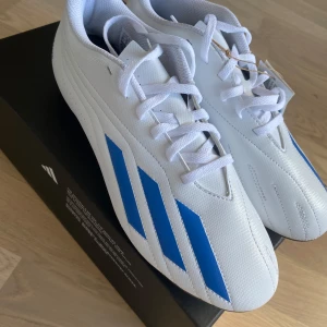 Vita fotbollsskor från Adidas - Snygga vita fotbollsskor från Adidas med blå detaljer och snörning. Perfekta för fotbollsspelare som vill ha stil och funktionalitet. Passar även som 41,5