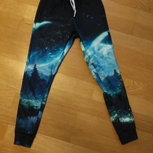 Mjukisbyxor med norrskensmönster från Mr. Gugu & Miss Go - Snygga mjukisbyxor med ett fantastiskt norrskensmönster i blå och svarta nyanser. Byxorna har en elastisk midja med vit snörning och är perfekta för en avslappnad stil. De är tillverkade i ett mjukt och bekvämt material. Helt oanvända 