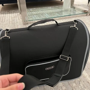 Svart transportbur med axelrem - Snygg svart transportbur med justerbar axelrem och dragkedja. Väskan har en rymlig insida med nätfönster och en ytterficka för extra förvaring. Perfekt för att bära dina hundar, katter, kaniner osv. Den är precis som ny, inga skador. Använd ett par gånger bara! Otrolig bra och stor bur!