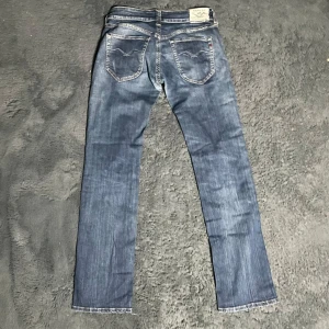 Blå jeans från Replay - Snygga blå jeans från Replay med klassisk femficksdesign och diskreta slitningar. De har en rak passform och är perfekta för en avslappnad stil. Märkeslappen syns tydligt på baksidan. 