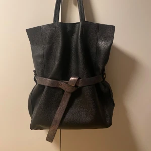Svart axelväska med bälte - Snygg svart totebag i skinn med ett dekorativt bälte runtom. Väskan har en stilren design med två handtag för enkel bärning.