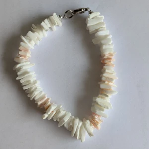 Vitt armband med snäckdetaljer - Charmigt armband med vita och ljusrosa snäckbitar. Armbandet har en praktisk metallåsning och ger en naturlig och bohemisk känsla. Perfekt för att lägga till en touch av strandvibbar till din stil.