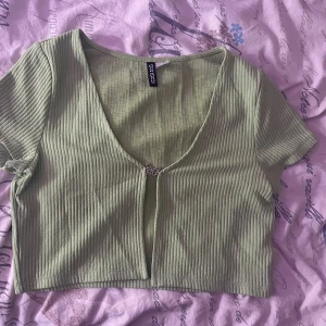 Grön ribbad kortärmad topp från HM - Säljer en grön ribbad topp från HM med korta ärmar och en djup v-ringning. Toppen är croppad och har en dekorativ knäppning framtill. Perfekt för en avslappnad stil.