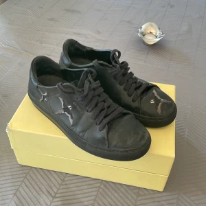 Svarta Axel Arigatos sneakers - Snygga svarta sneakers från Axel Arigato med unikt fågelmotiv. Skorna har snörning och är tillverkade i skinn, vilket ger en lyxig känsla. Perfekta för dig som vill ha en stilren men ändå iögonfallande look. Lådan medföljer!!