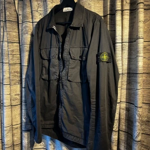 Svart overshirt från Stone Island - Säljer en stilren svart overshirt från Stone Island med dragkedja och två bröstfickor med knappar. Jackan har en ikonisk logotyp på ärmen och är i storlek L