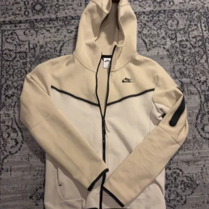 Beige hoodie från Nike - Säljer en snygg beige hoodie från Nike med dragkedja och svart logga på bröstet. Den har en praktisk ficka med dragkedja på ärmen och en justerbar huva. Perfekt för en sportig look! Skick 7/10 och är knappast använd, frågor? DM! pris kan diskuteras vid snabb affär!