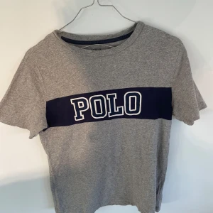 Grå t-shirt från Ralph Lauren - Säljer en grå t-shirt från Ralph Lauren med en stor mörkblå rand över bröstet där det står 'POLO' i vita bokstäver. T-shirten är kortärmad och gjord i bomull, perfekt för en avslappnad stil. Säljer en liknade artikel så kika gärna in den?😊