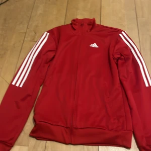 Röd träningsjacka från Adidas - Säljer en röd träningsjacka från Adidas med klassiska vita ränder längs ärmarna och dragkedja framtill. Perfekt för sportiga aktiviteter eller som en stilren vardagsjacka.